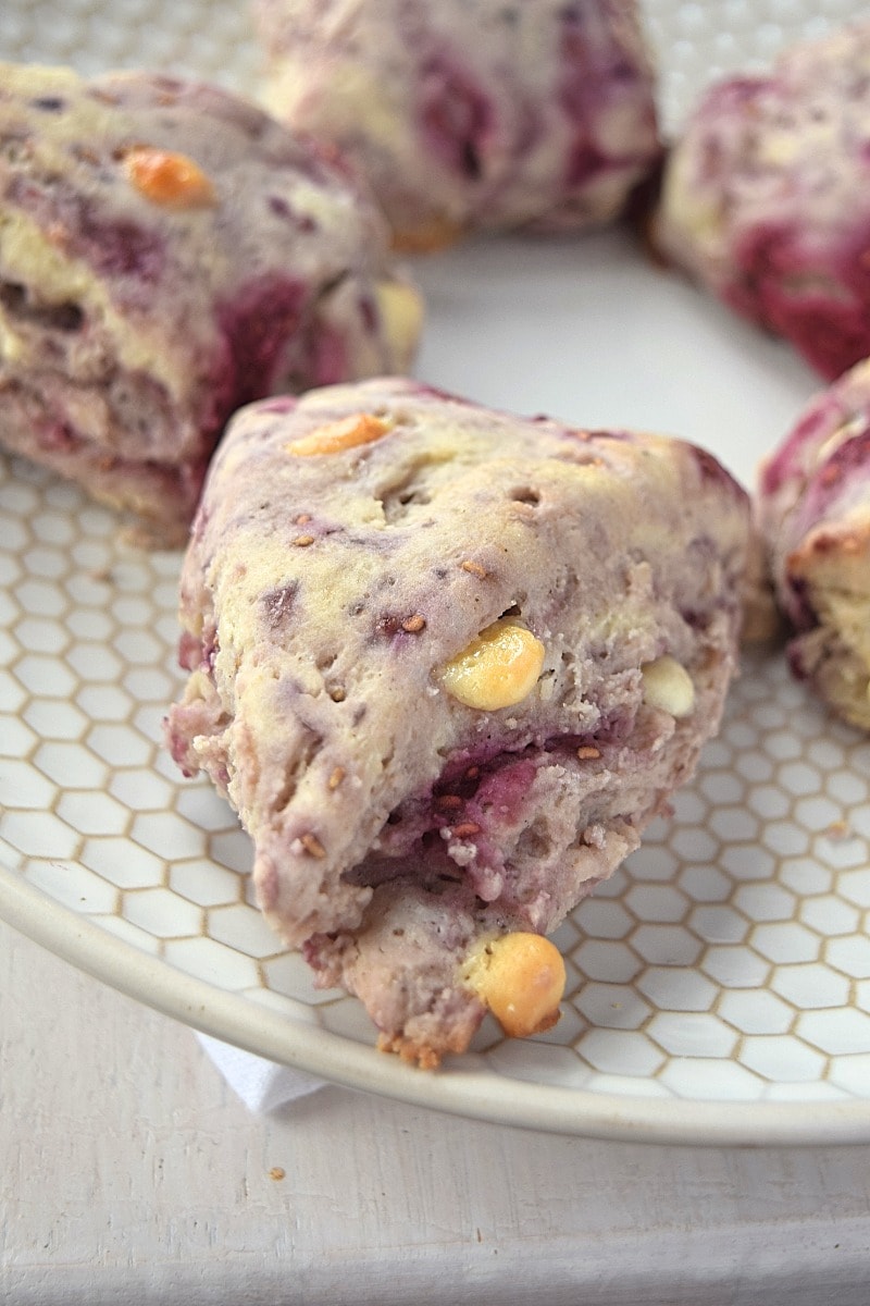 A plate of gluten free raspberry white chocolate scones. Featuring one in the front and a few in the background.