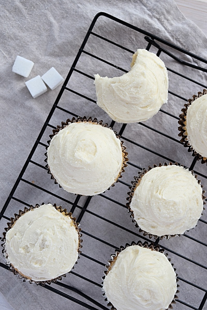 A top-down view of gluten free vanilla cupcakes on a baking rack. There's a bite taken out of one of them. 